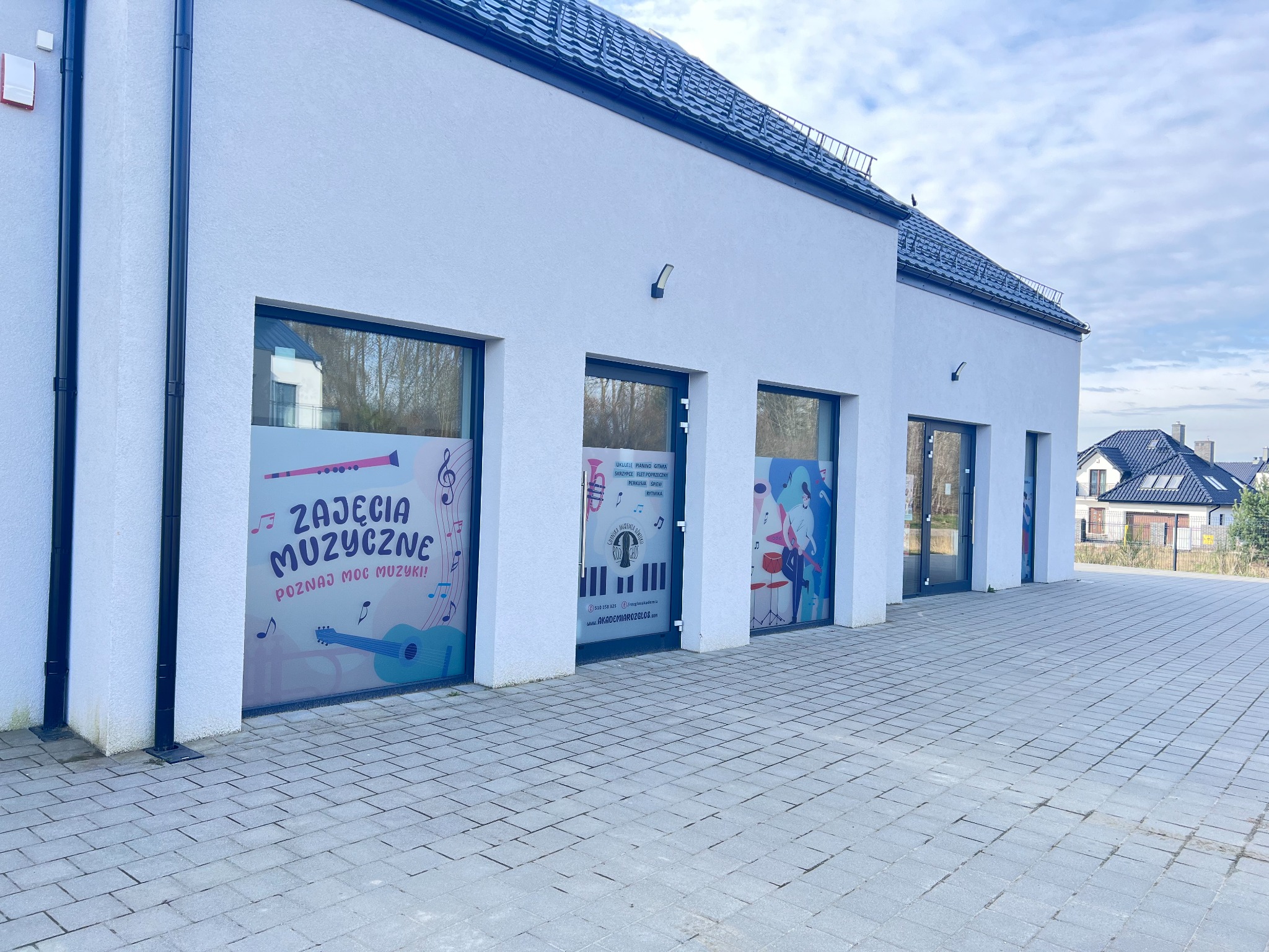 Nowoczesny budynek z oknami i naklejkami reklamującymi zajęcia muzyczne, w tym ukulele, pianino, gitarę, skrzypce, flet poprzeczny, perkusję, śpiew i rytmikę.