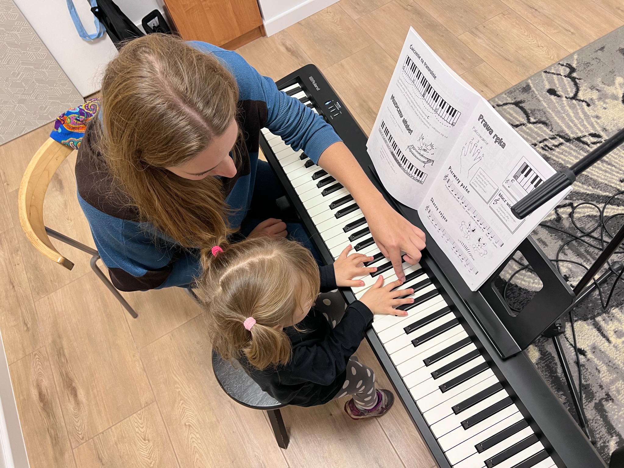 Widok z góry: Kobieta z długimi włosami uczy małą dziewczynkę grać na keyboardzie, wskazując palcem na klawisze. Na statywie nuty z ćwiczeniami i alfabetem muzycznym.