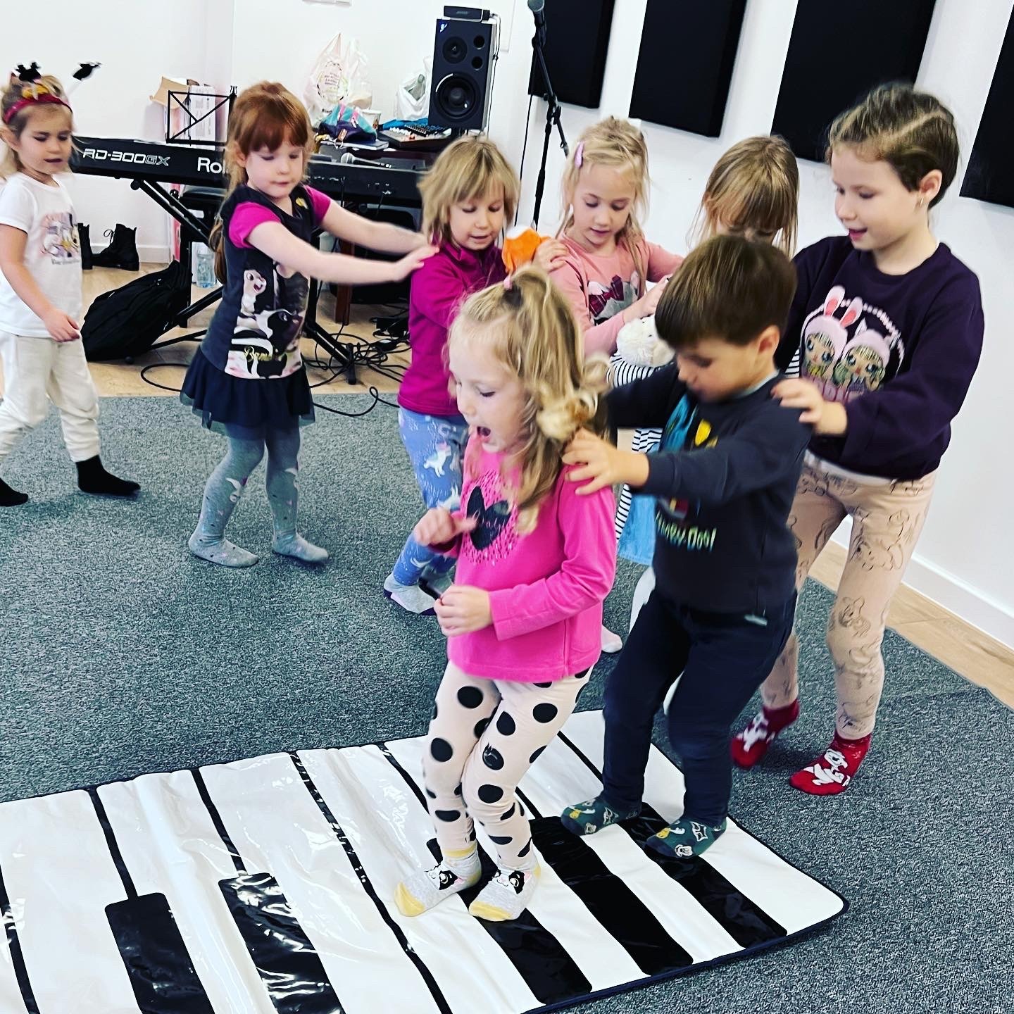 Grupa dzieci bawi się na macie imitującej klawiaturę fortepianu w jasnym pomieszczeniu z pianinem i panelami akustycznymi na ścianie.