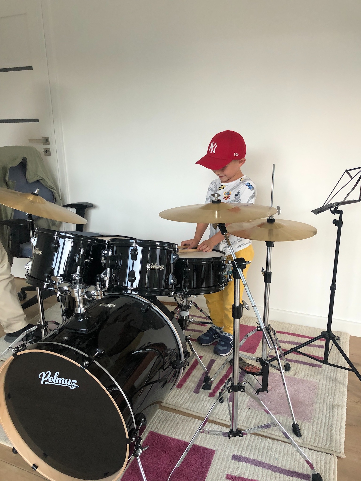 Chłopiec w czerwonej czapce New York Yankees uczy się grać na czarnej perkusji Polmuz w jasnym pokoju, nuty na pulpicie obok.
