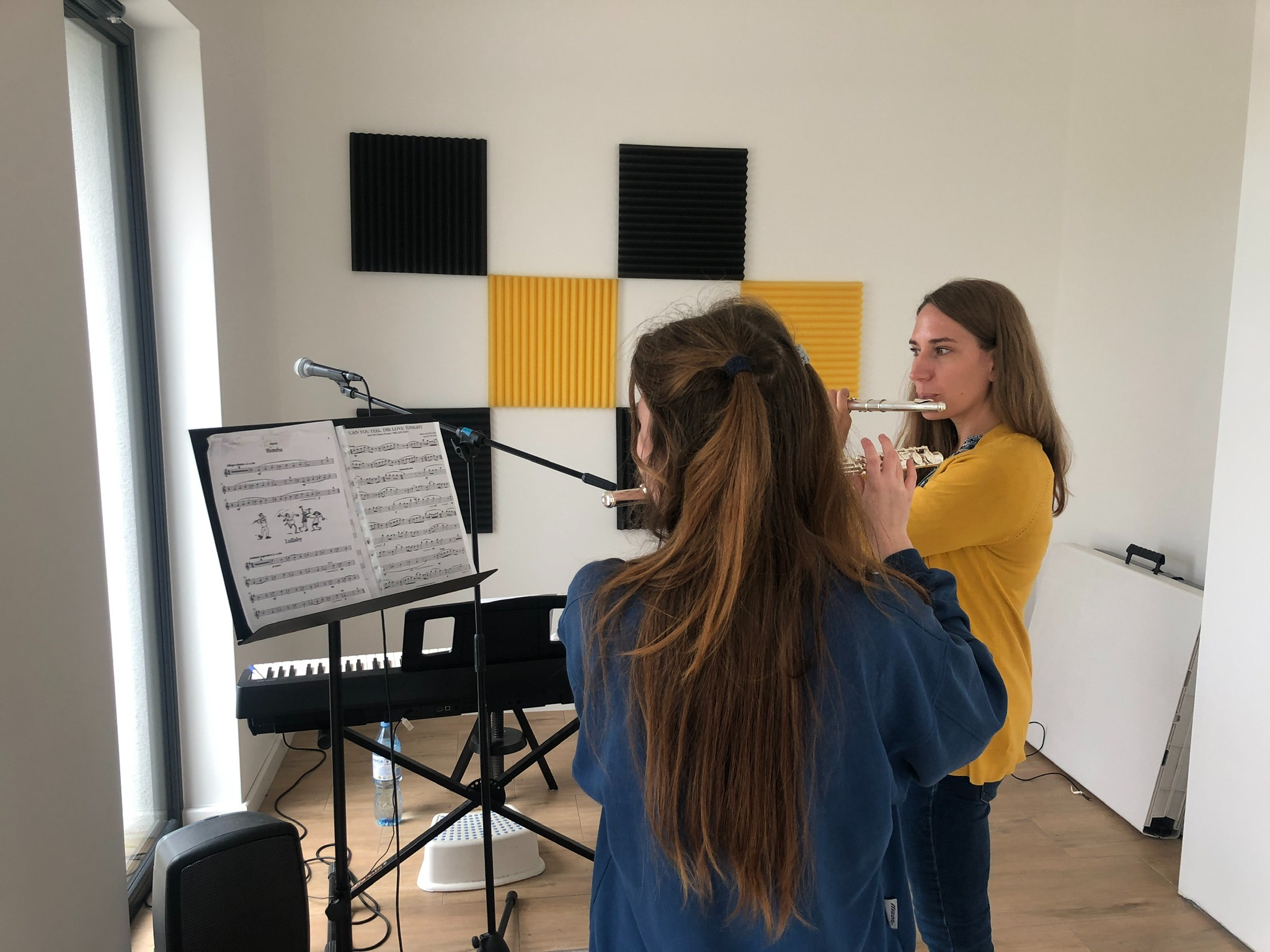 Dwie młode kobiety ćwiczą grę na fletach w pokoju z panelami akustycznymi na ścianie, nutami na pulpicie i keyboardem w tle.