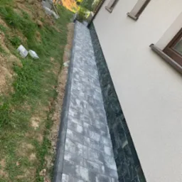 Ukończony chodnik z szarej kostki brukowej biegnący wzdłuż ściany domu z oknami, z widocznym fragmentem trawnika i stertą kostki na zboczu.