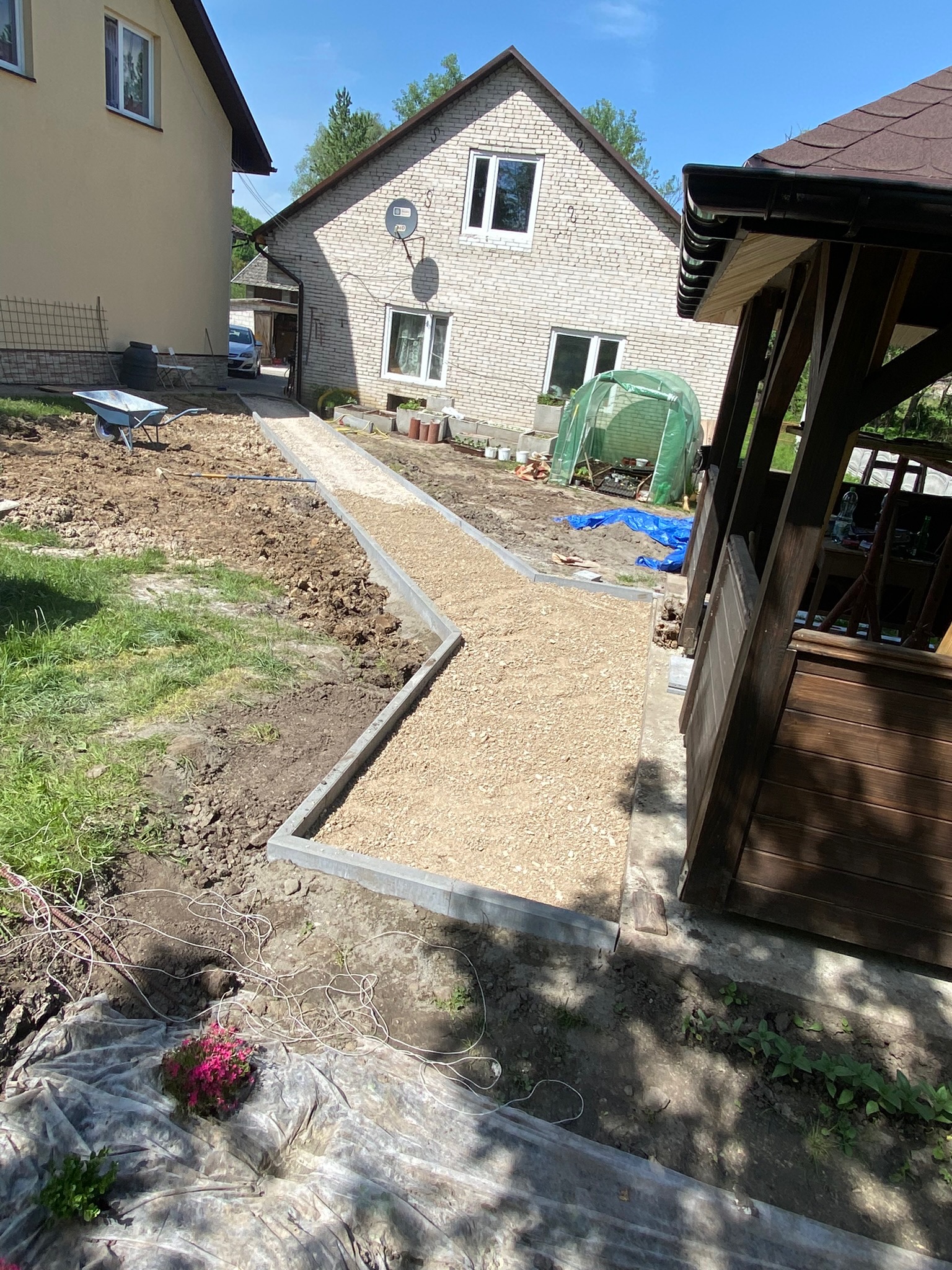 Budowa ścieżki ogrodowej z obrzeżami i podsypką żwirową, widoczny wózek budowlany, dom i altana w tle, częściowo rozplantowana ziemia.