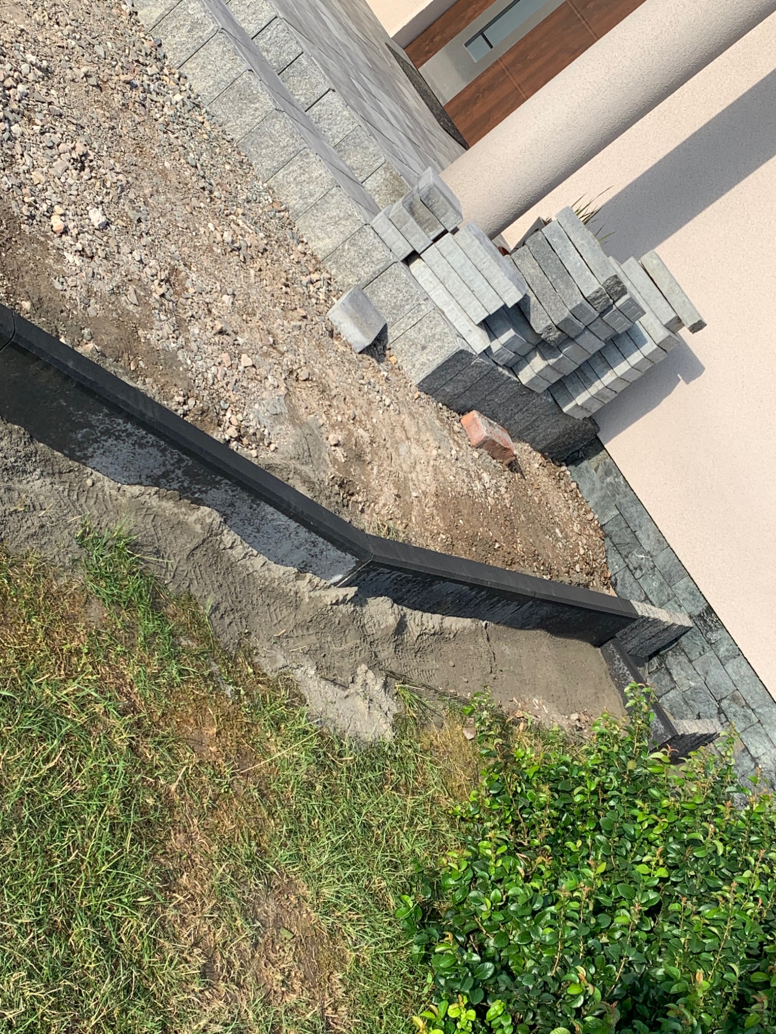 Przygotowanie podłoża pod ułożenie kostki brukowej: widoczne krawężniki, podsypka z kruszywa, stos kostki oraz fragment trawnika.