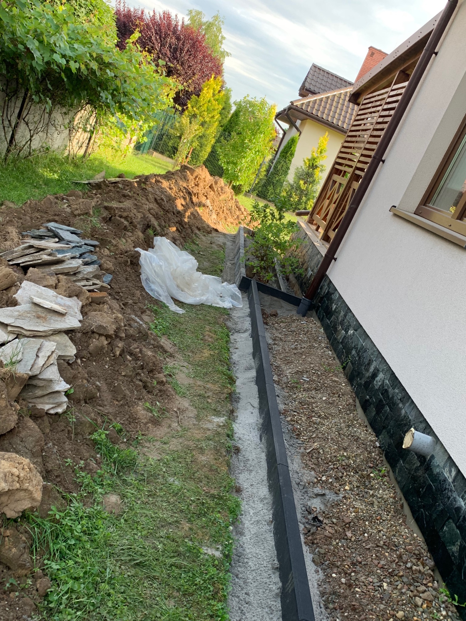 Przygotowanie terenu przy budynku: wykop z betonową krawędzią, podsypka żwirowa, sterta ziemi i kamieni w ogrodzie.