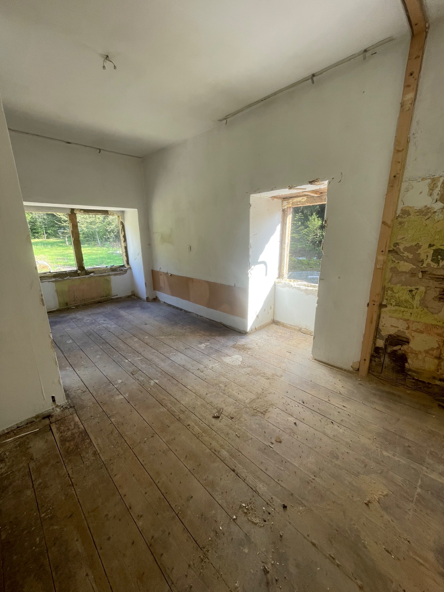 Wnętrze pokoju w Jeleniej Górze przed remontem: surowe drewniane podłogi, ściany z resztkami tapet, okna bez ram z widokiem na zieleń.