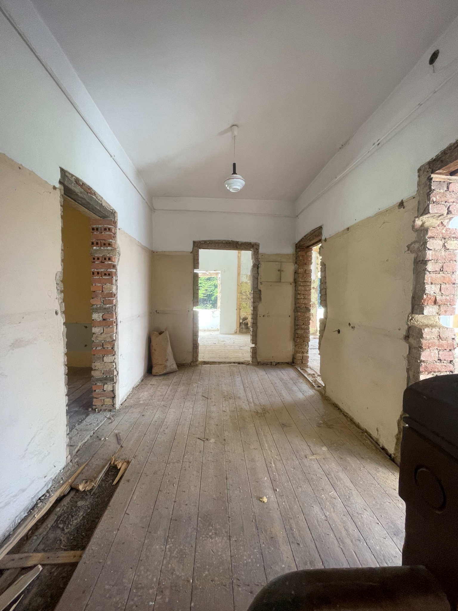 Wnętrze mieszkania w trakcie demontażu: odsłonięte cegły przy otworach drzwiowych, drewniana podłoga z ubytkami, worki z gruzem w tle, widok perspektywiczny przez kolejne pomieszczenia.