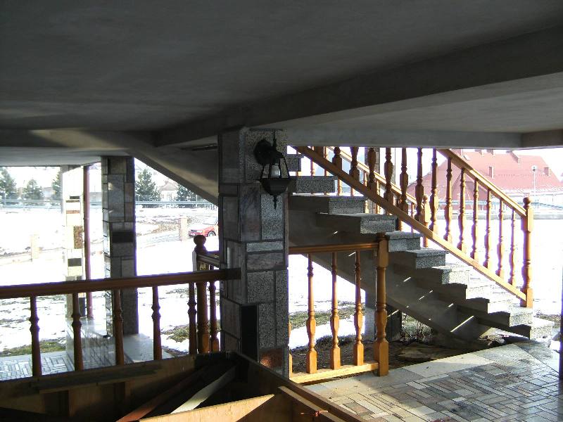 Wnętrze z betonowymi schodami z drewnianą balustradą i kamiennymi kolumnami, widok na zewnątrz w zimowy dzień.