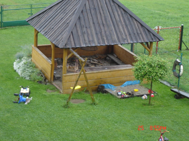 Drewniana piaskownica z zadaszeniem i huśtawką na trawniku, obok mniejsza piaskownica z zabawkami, w tle ogrodzenie.
