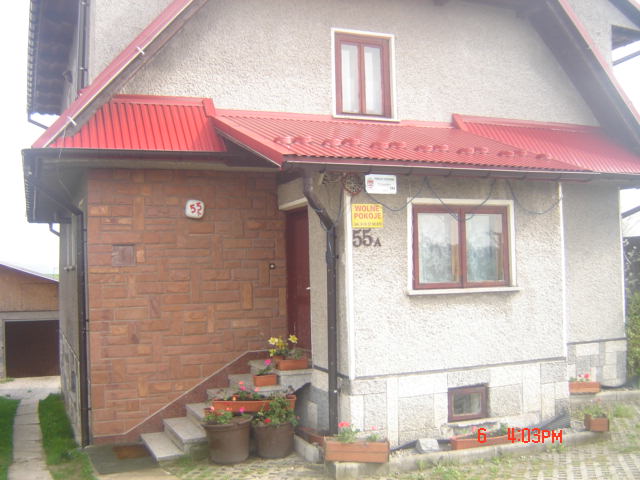Dom z czerwonym dachem i numerem 55A, tabliczka z napisem 'Wolne Pokoje', kwiaty w donicach na schodach.