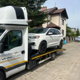 Biały samochód osobowy Ford Edge z czarnymi felgami, zabezpieczony pasami transportowymi, załadowany na lawetę pomocy drogowej firmy EWINCAR na tle brukowanej ulicy i domów.
