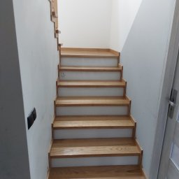 Stolarnia Babice - Jasne, dębowe stopnie schodów z białymi podstopnicami, widziane od dołu, z fragmentem ażurowej balustrady na górze i szarą ścianą po lewej stronie.