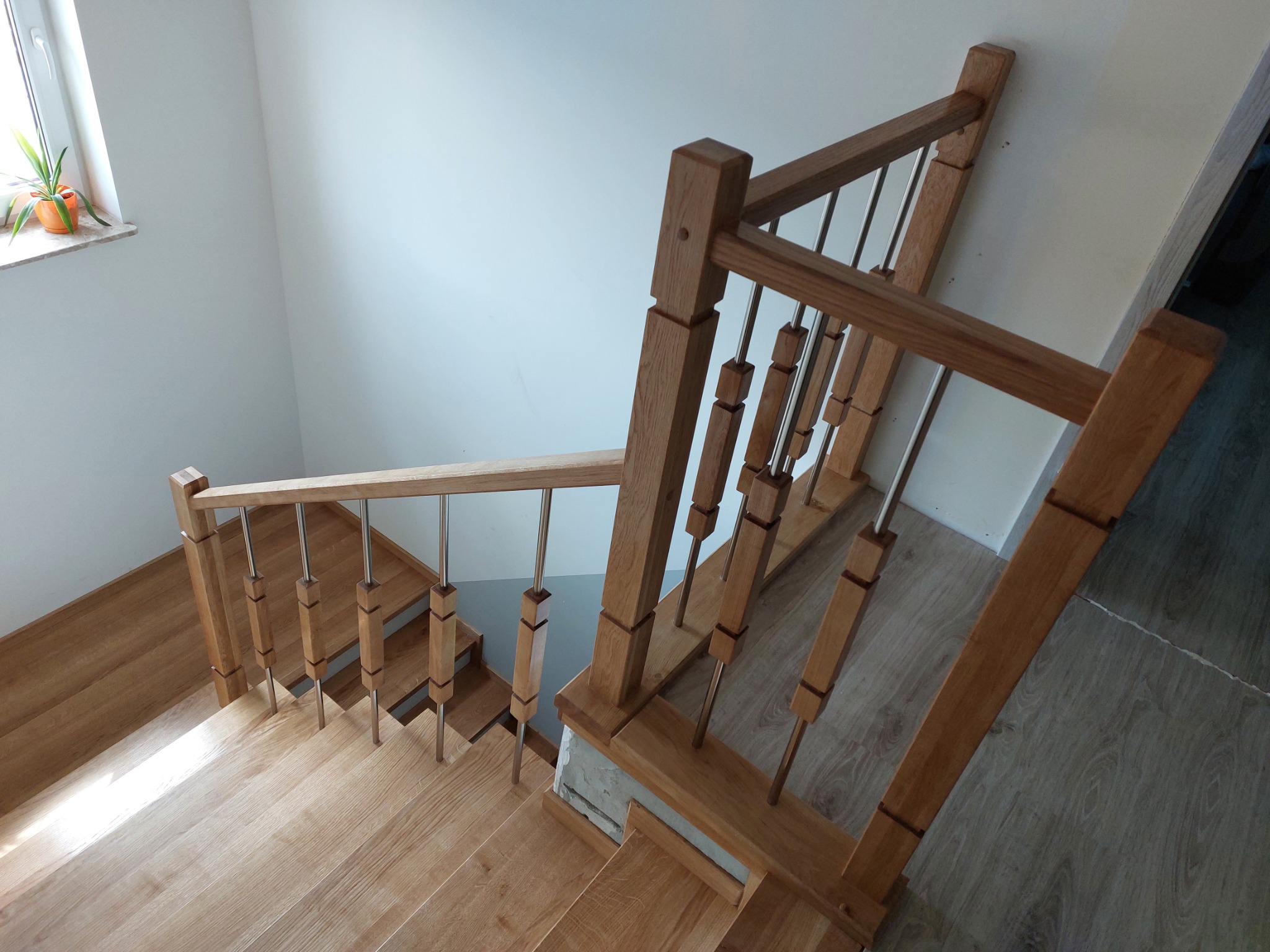 Wysoki kąt widzenia na jasne, drewniane schody z metalowymi tralkami i drewnianą balustradą w nowoczesnym wnętrzu, widoczny parapet z rośliną doniczkową.