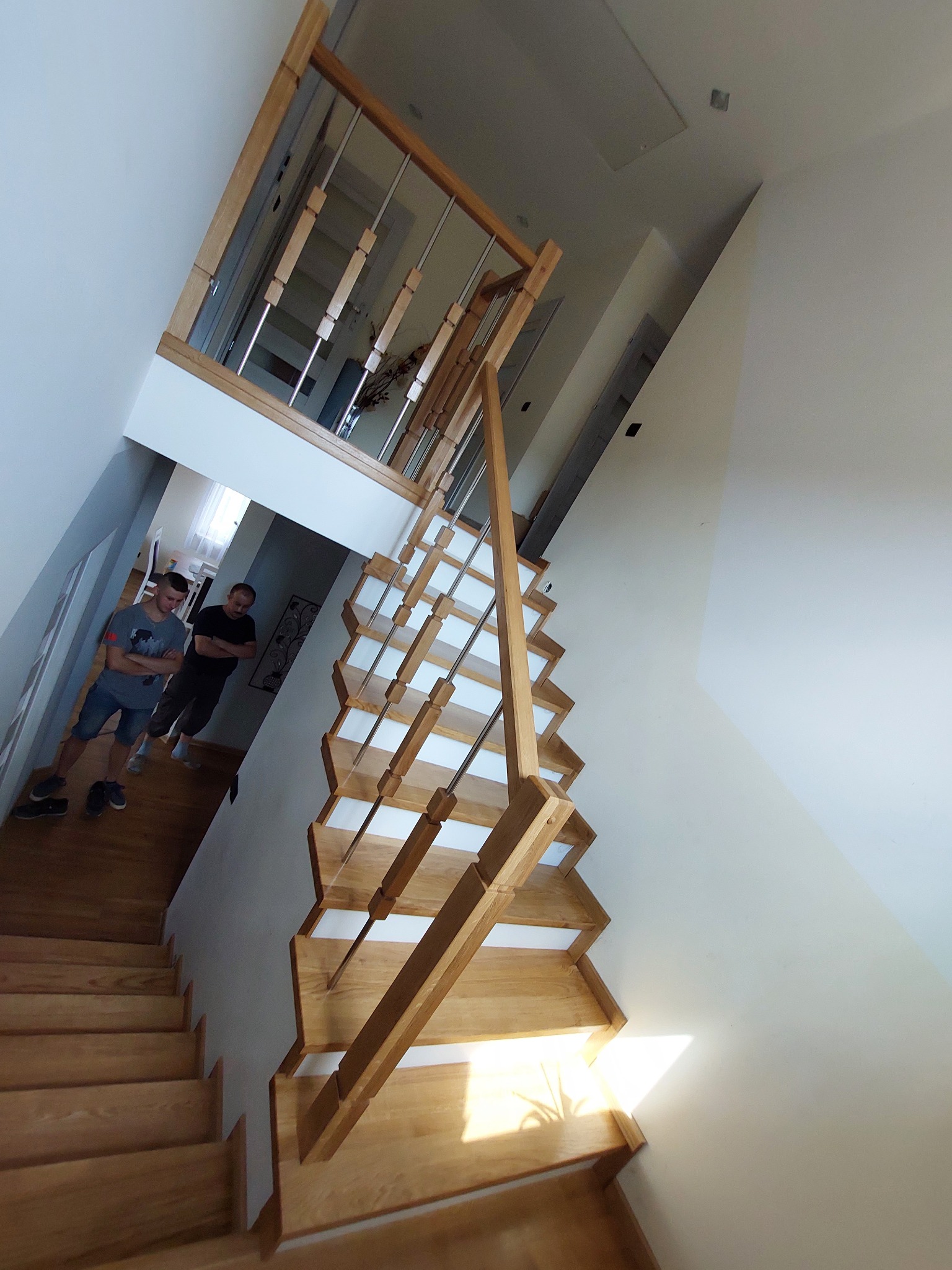 Widok z góry na jasne, drewniane schody z metalowymi balustradami w nowoczesnym wnętrzu, z dwoma mężczyznami stojącymi na dole schodów.