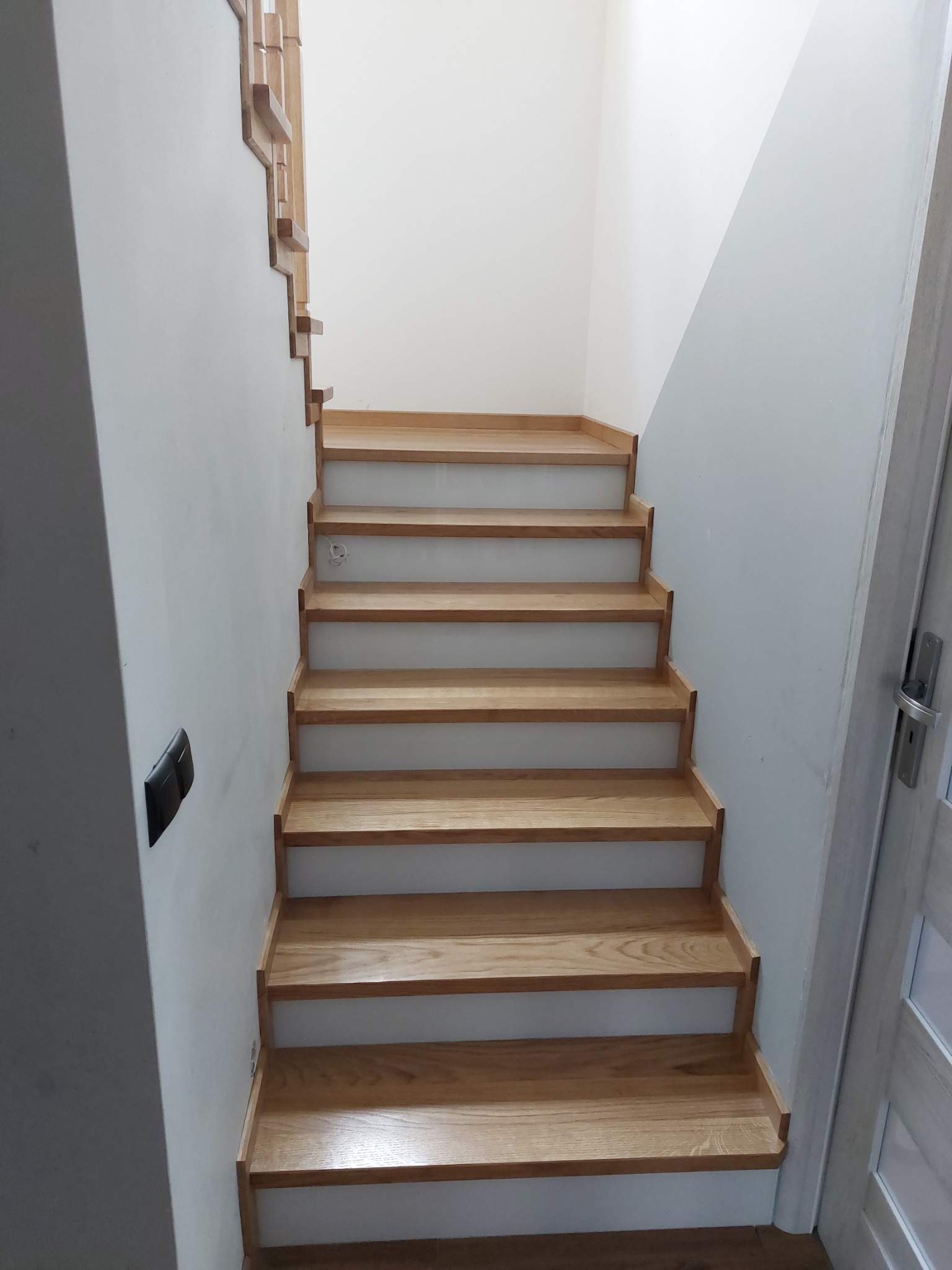 Jasne, dębowe stopnie schodów z białymi podstopnicami, widziane od dołu, z fragmentem ażurowej balustrady na górze i szarą ścianą po lewej stronie.