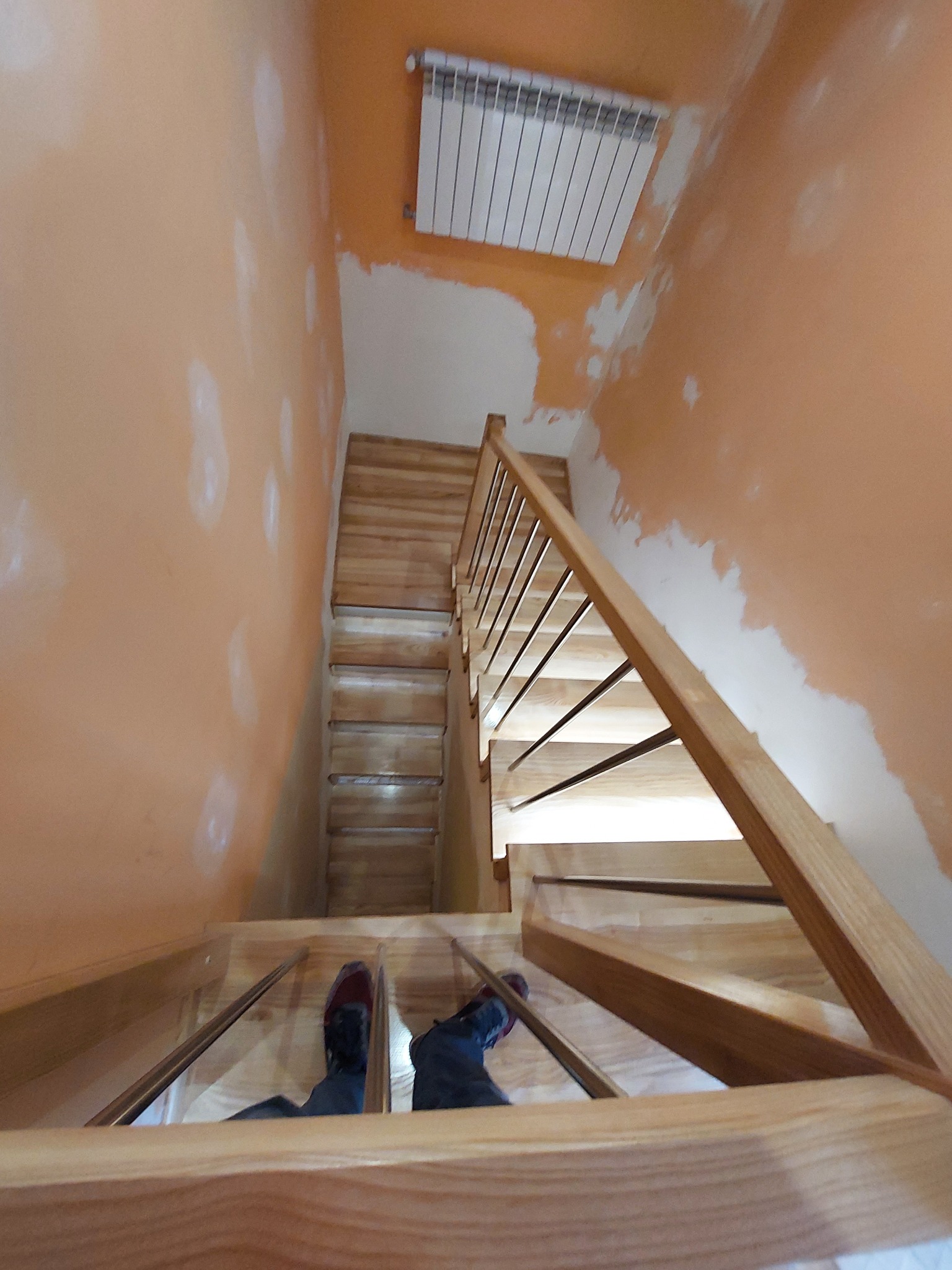 Widok z góry na drewniane schody z metalowymi balustradami w trakcie montażu, ściany pomalowane na pomarańczowo z widocznymi niedociągnięciami, buty osoby robiącej zdjęcie widoczne na pierwszym...