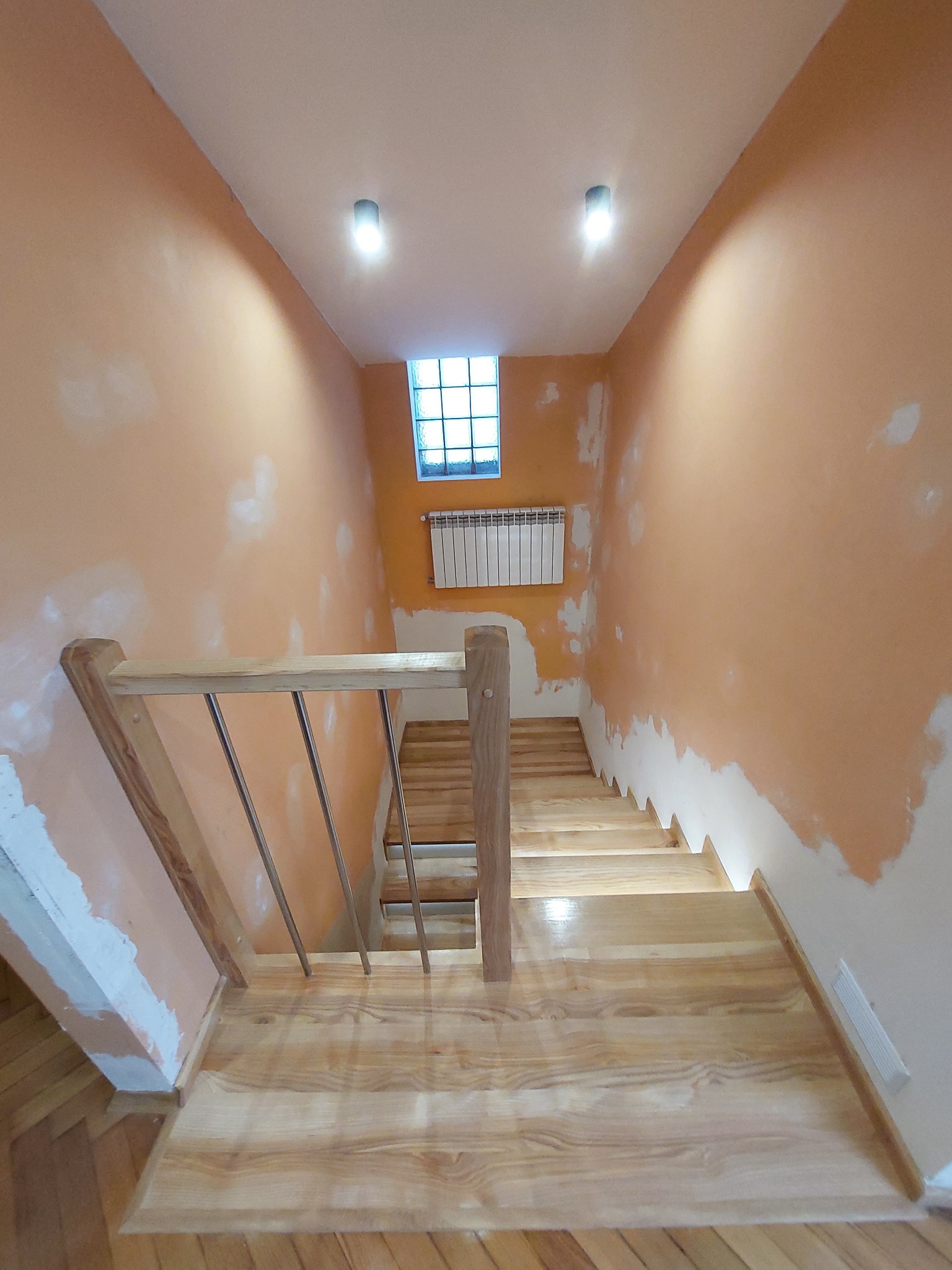 Wnętrze w trakcie wykańczania: jasne, drewniane schody z metalową balustradą w pomarańczowym pomieszczeniu, ściany z widocznymi śladami po gładzi, małe okno z luksferami i grzejnik na ścianie.