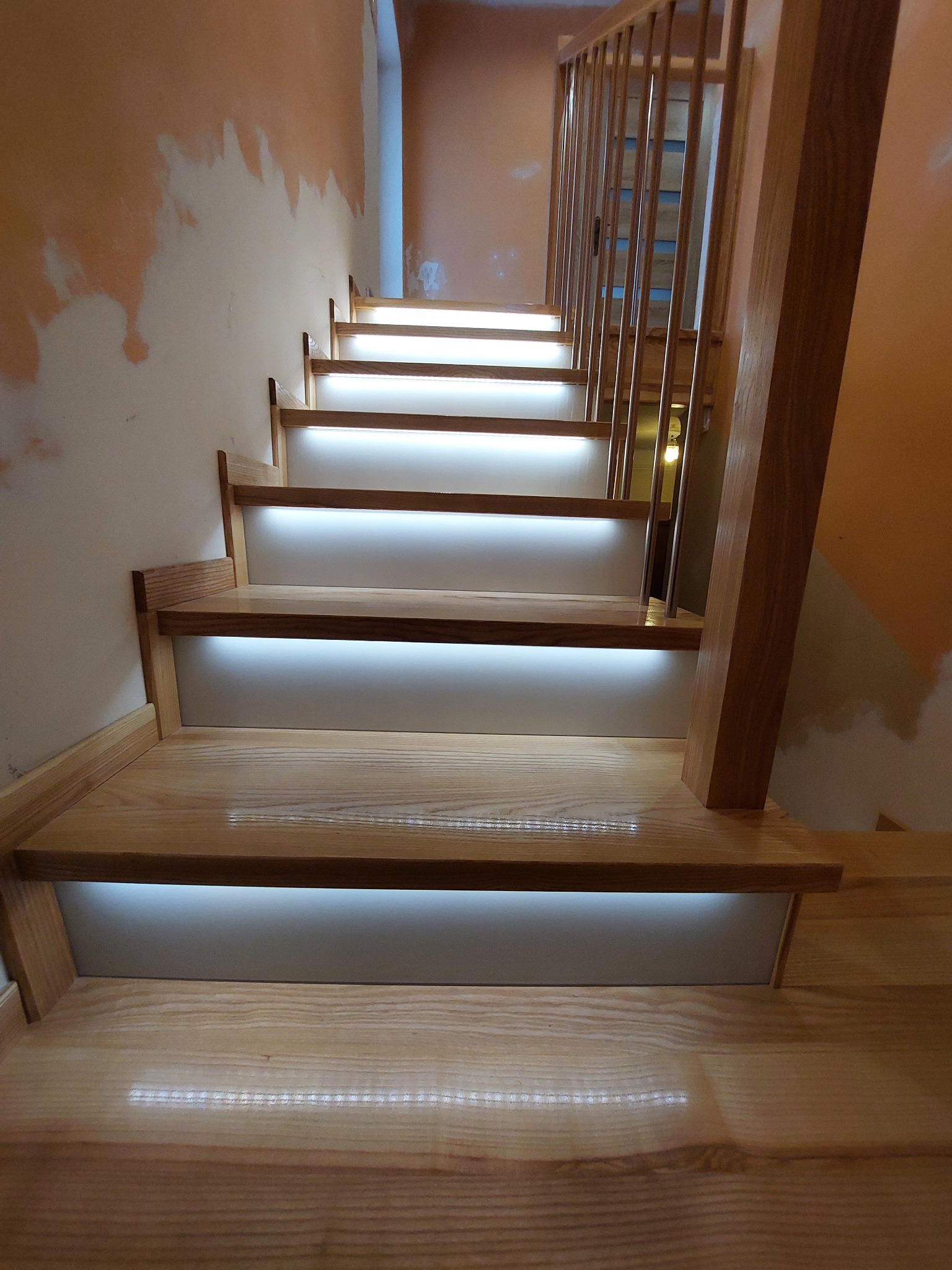 Jasne drewniane schody z podświetleniem LED na każdym stopniu, prowadzące do góry, z widoczną drewnianą balustradą i niedokończonym malowaniem ścian w tle.