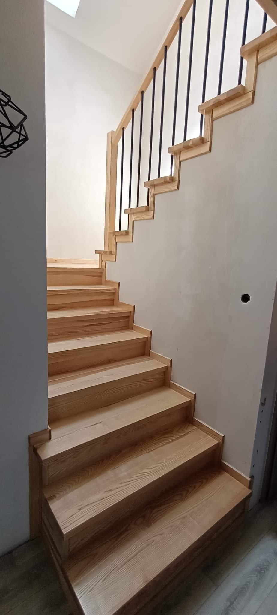 Jasne, drewniane schody z czarnymi metalowymi prętami balustrady, widok od dołu, oświetlone z góry