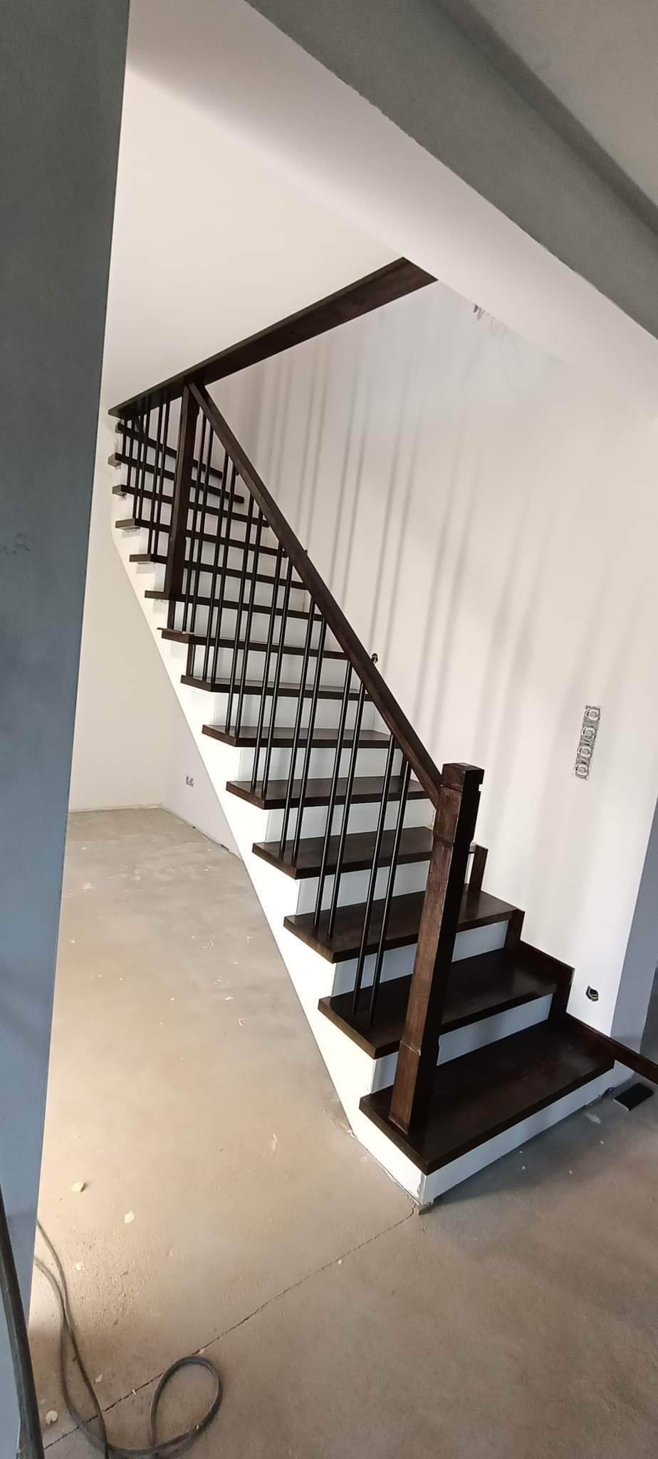 Schody drewniane z ciemnymi stopniami i balustradą z czarnymi prętami, widoczne w surowym stanie wykończenia wnętrza.