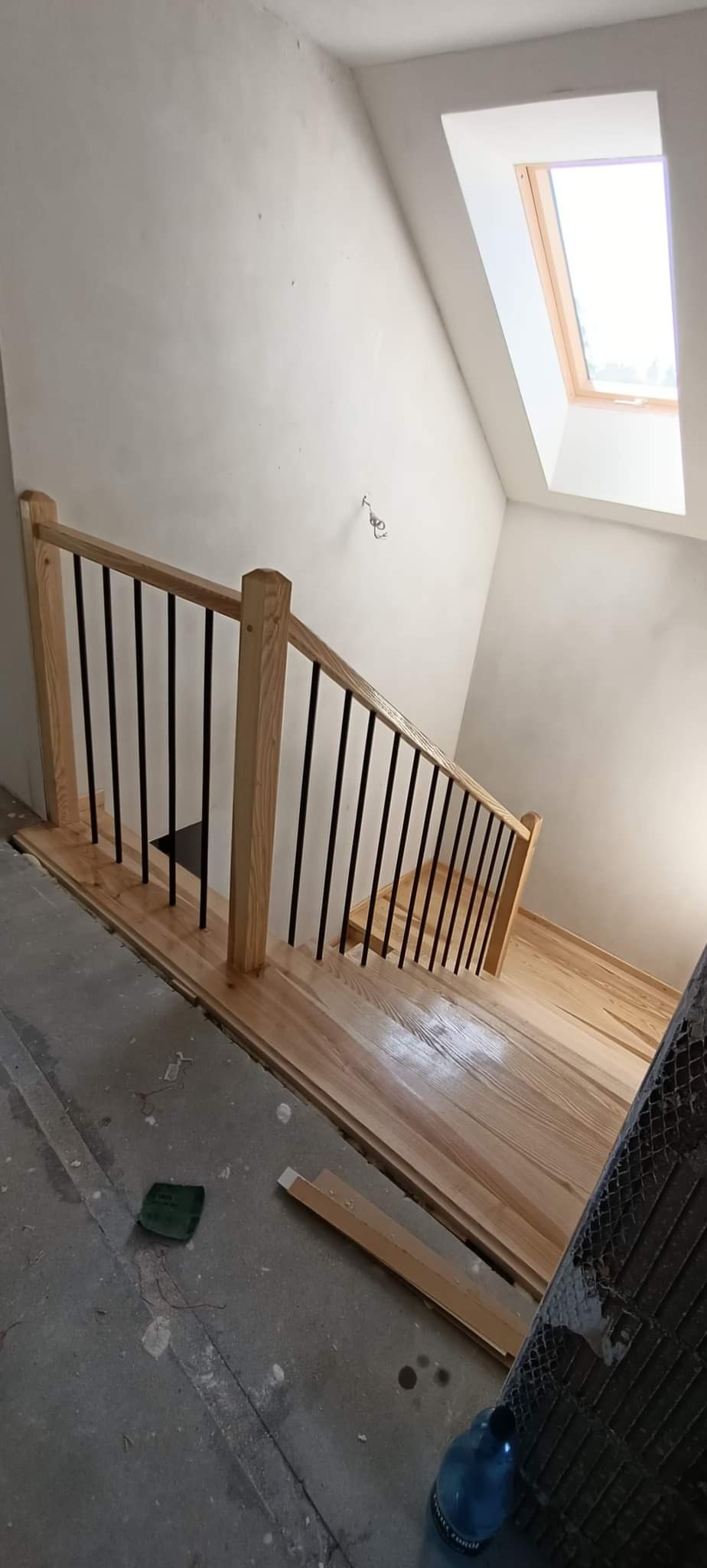 Wnętrze w trakcie remontu z widokiem na drewniane schody z metalową balustradą i oknem dachowym.