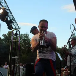 Mężczyzna grający na trąbce na scenie podczas występu plenerowego, widoczne oświetlenie sceniczne i instrumenty pozostałych muzyków.