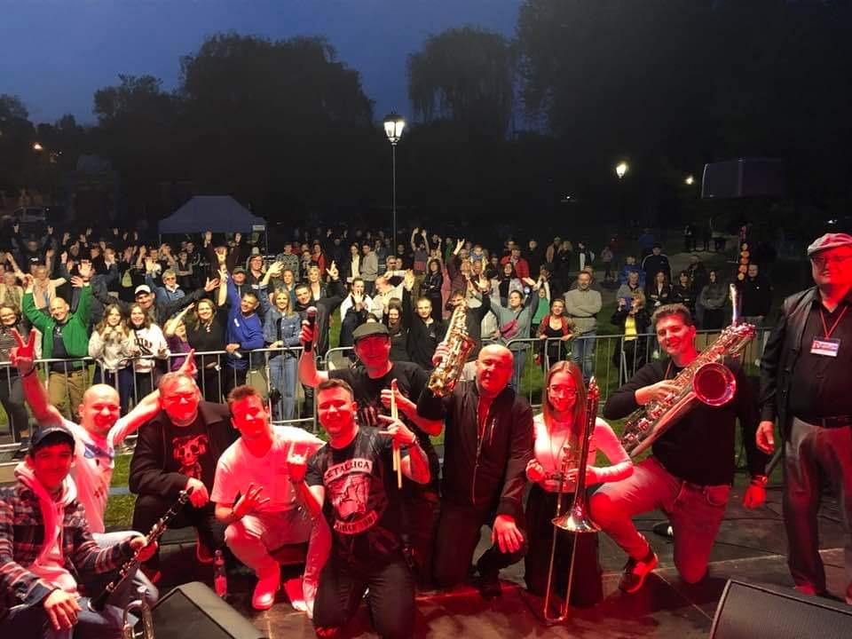 Zespół muzyczny na scenie podczas koncertu plenerowego, widoczny tłum publiczności w tle, saksofony i puzon w rękach muzyków, koszulka z logo Metallica.