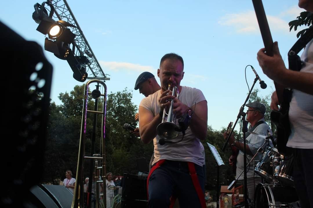 Mężczyzna grający na trąbce na scenie podczas występu plenerowego, widoczne oświetlenie sceniczne i instrumenty pozostałych muzyków.