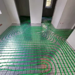 Wnętrze w trakcie instalacji ogrzewania podłogowego: zielone rury ułożone na folii z siatką, widoczne okna i surowy stan ścian.