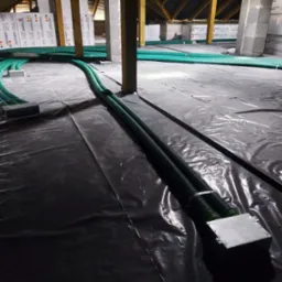 Zielone, karbowane rury wentylacyjne rozprowadzone na czarnej folii w przestrzeni poddasza z widoczną izolacją ze styropianu.