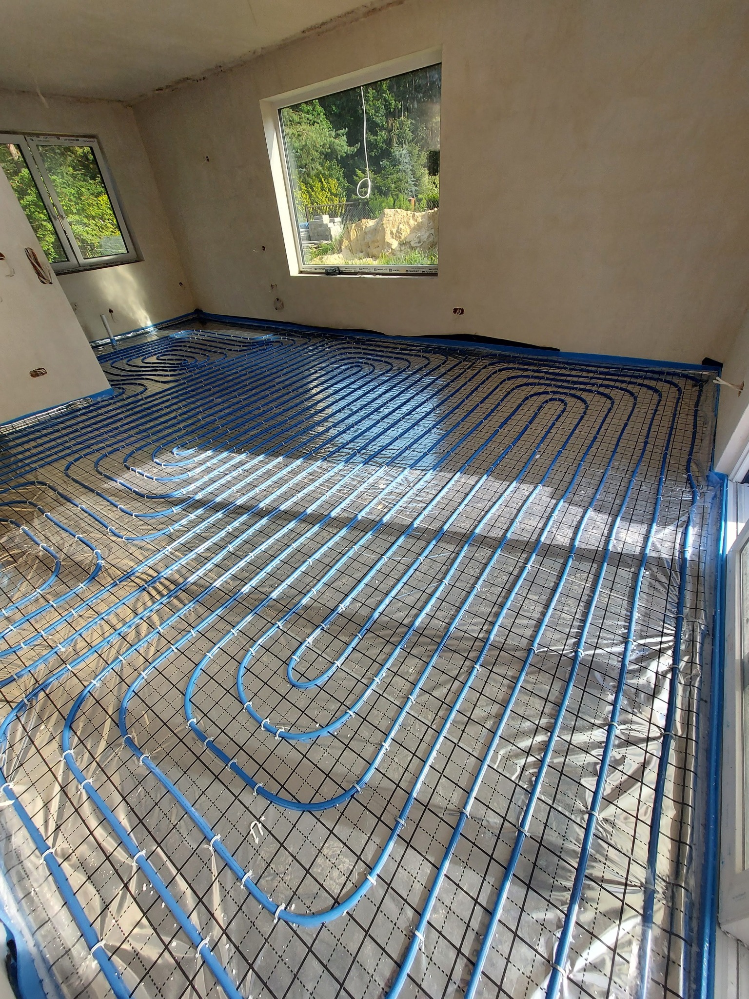 Niebieskie rury ogrzewania podłogowego ułożone na folii z siatką w surowym wnętrzu z dwoma oknami, widoczne promienie słoneczne.
