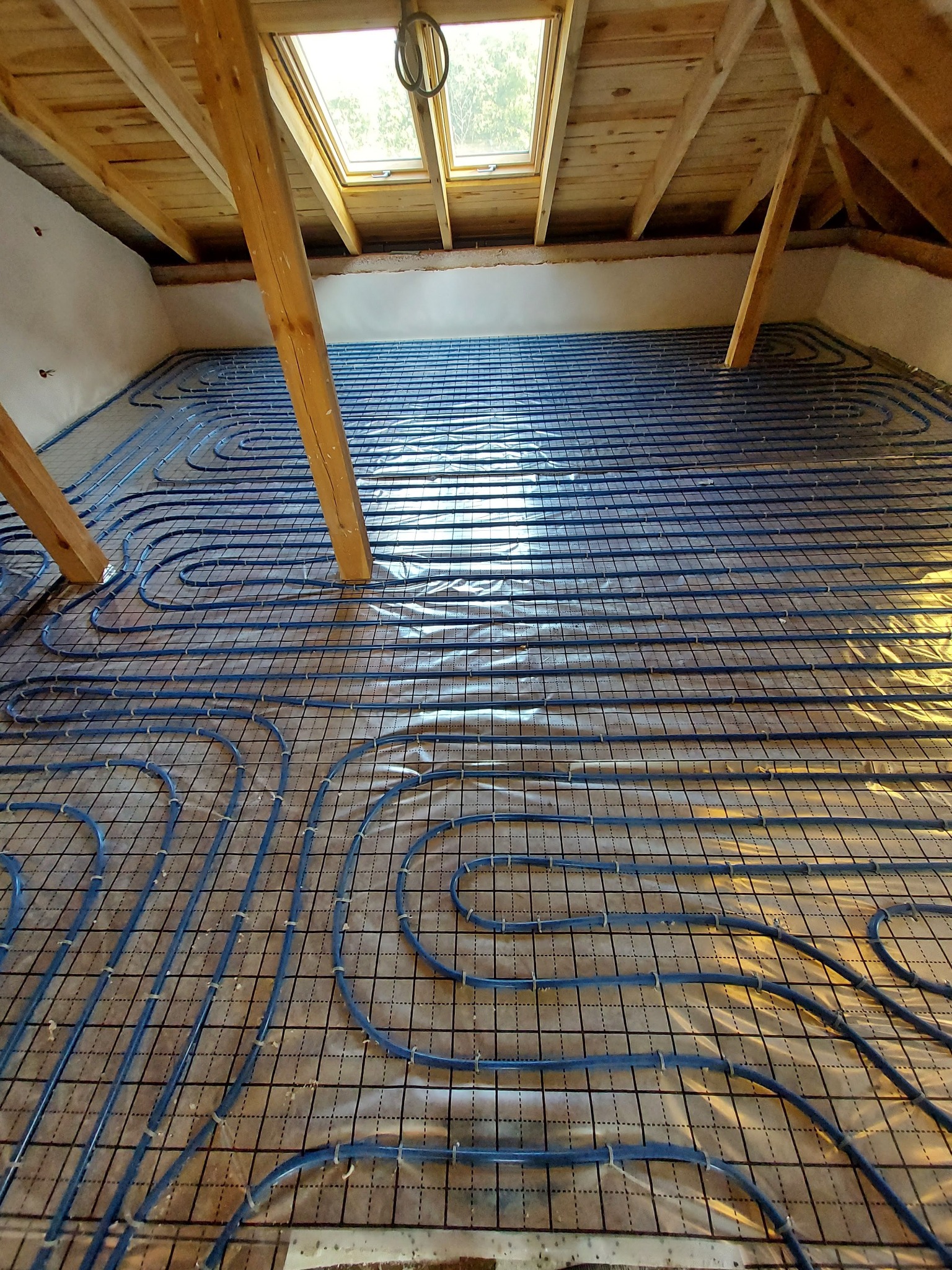 Montaż ogrzewania podłogowego w poddaszu z widoczną konstrukcją dachu i świetlikiem, niebieskie rurki rozłożone na folii i siatce.