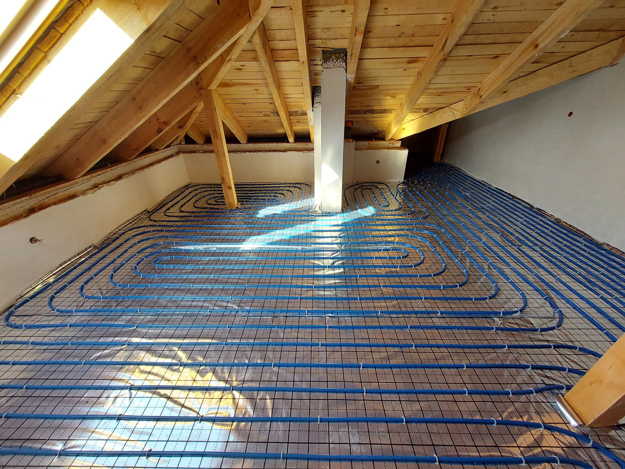 Niebieskie rurki ogrzewania podłogowego ułożone na metalowej siatce, widok z góry na poddaszu z drewnianą konstrukcją dachu i oknem dachowym.