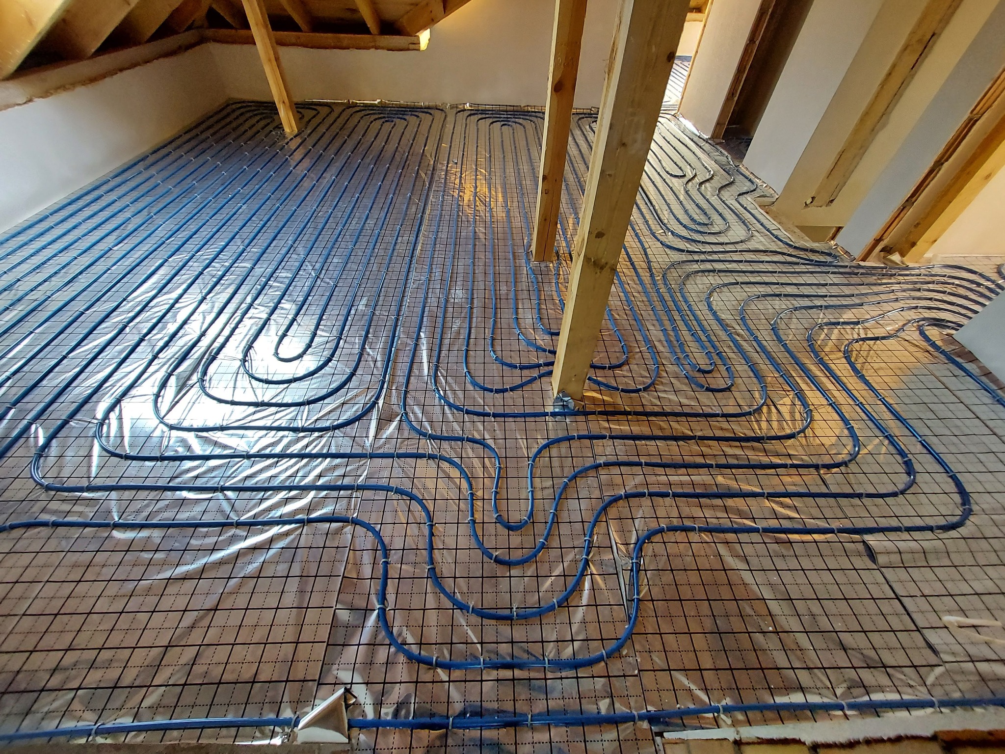 Niebieskie rury ogrzewania podłogowego ułożone na folii z siatką, widoczne drewniane elementy konstrukcji dachu.