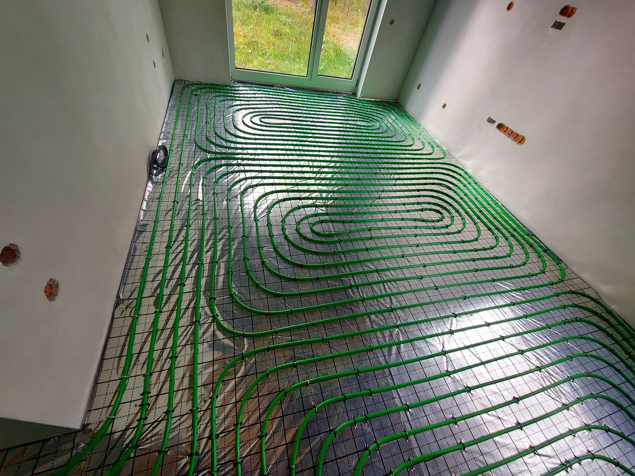 Zielone rury ogrzewania podłogowego ułożone spiralnie na folii izolacyjnej z siatką w surowym pomieszczeniu z oknem i widocznymi puszkami elektrycznymi w ścianie.