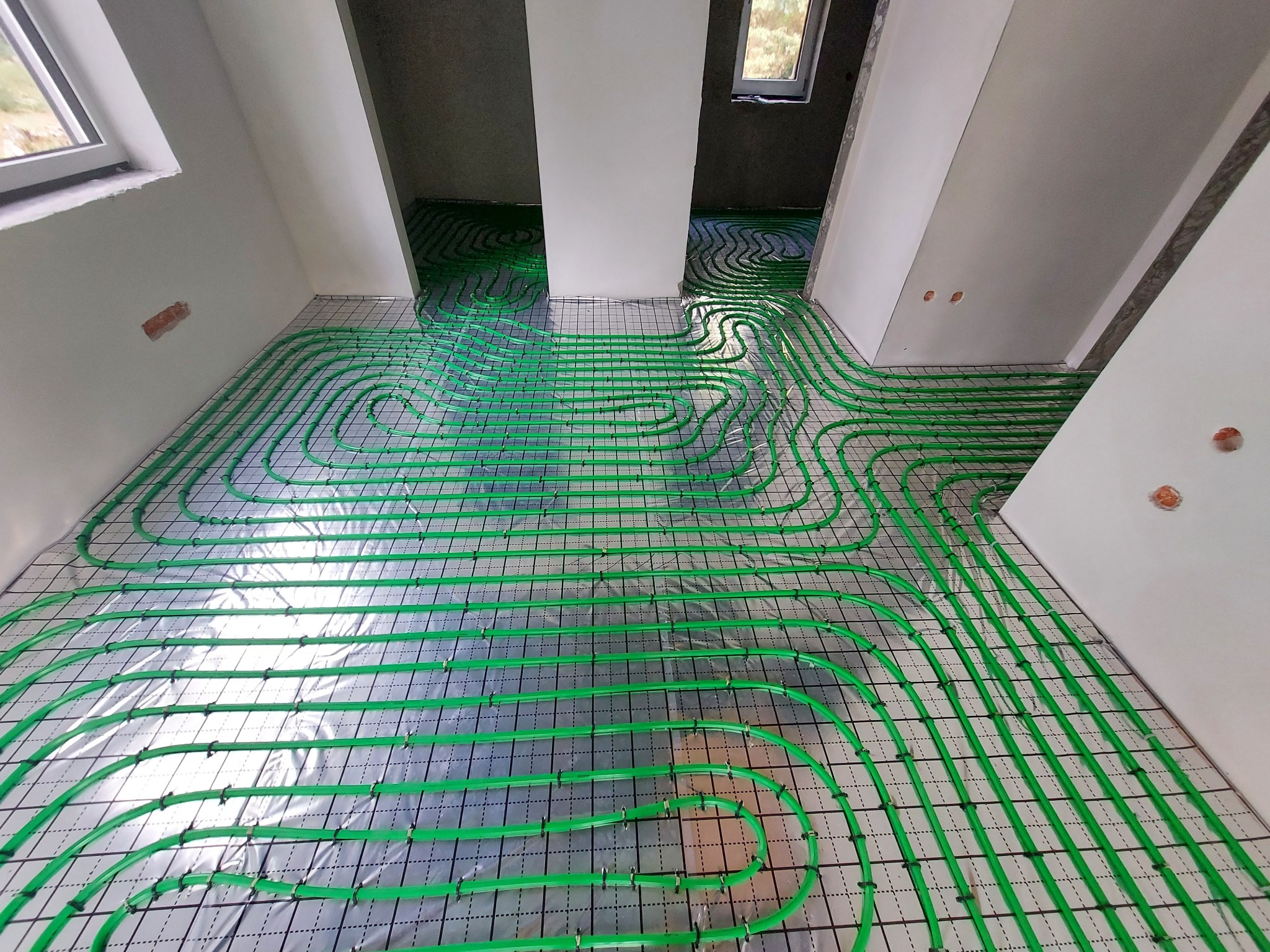 Wnętrze w trakcie instalacji ogrzewania podłogowego: zielone rury ułożone na folii z siatką, widoczne okna i surowy stan ścian.