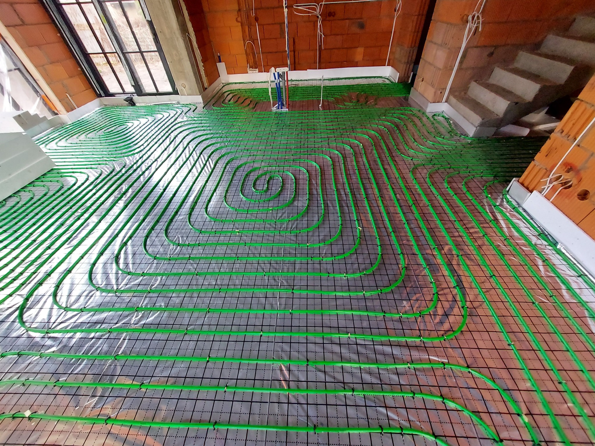 Zielone rury ogrzewania podłogowego ułożone spiralnie na folii izolacyjnej z siatką, widok z góry w surowym wnętrzu z ceglanymi ścianami i oknem.