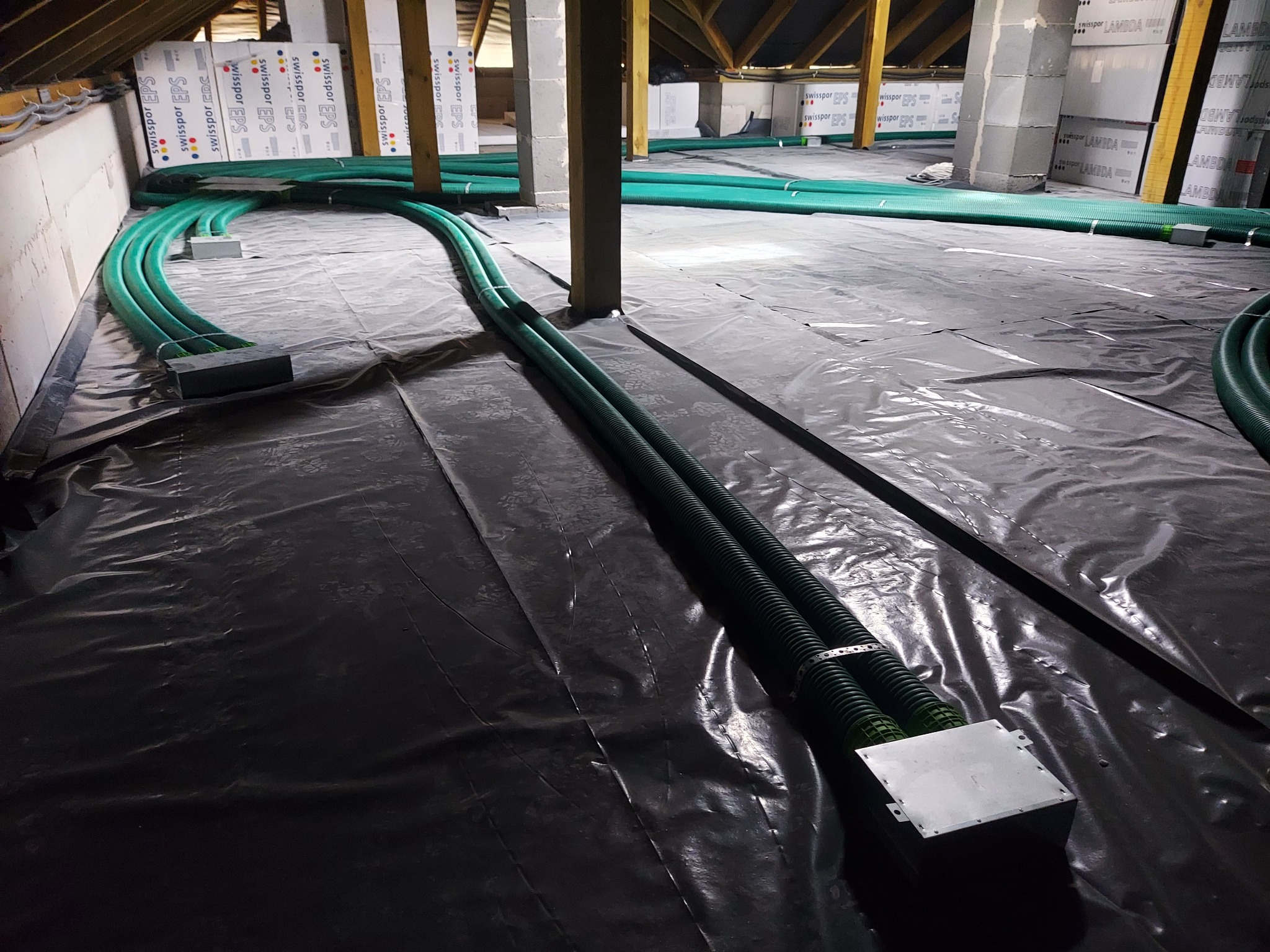 Zielone, karbowane rury wentylacyjne rozprowadzone na czarnej folii w przestrzeni poddasza z widoczną izolacją ze styropianu.