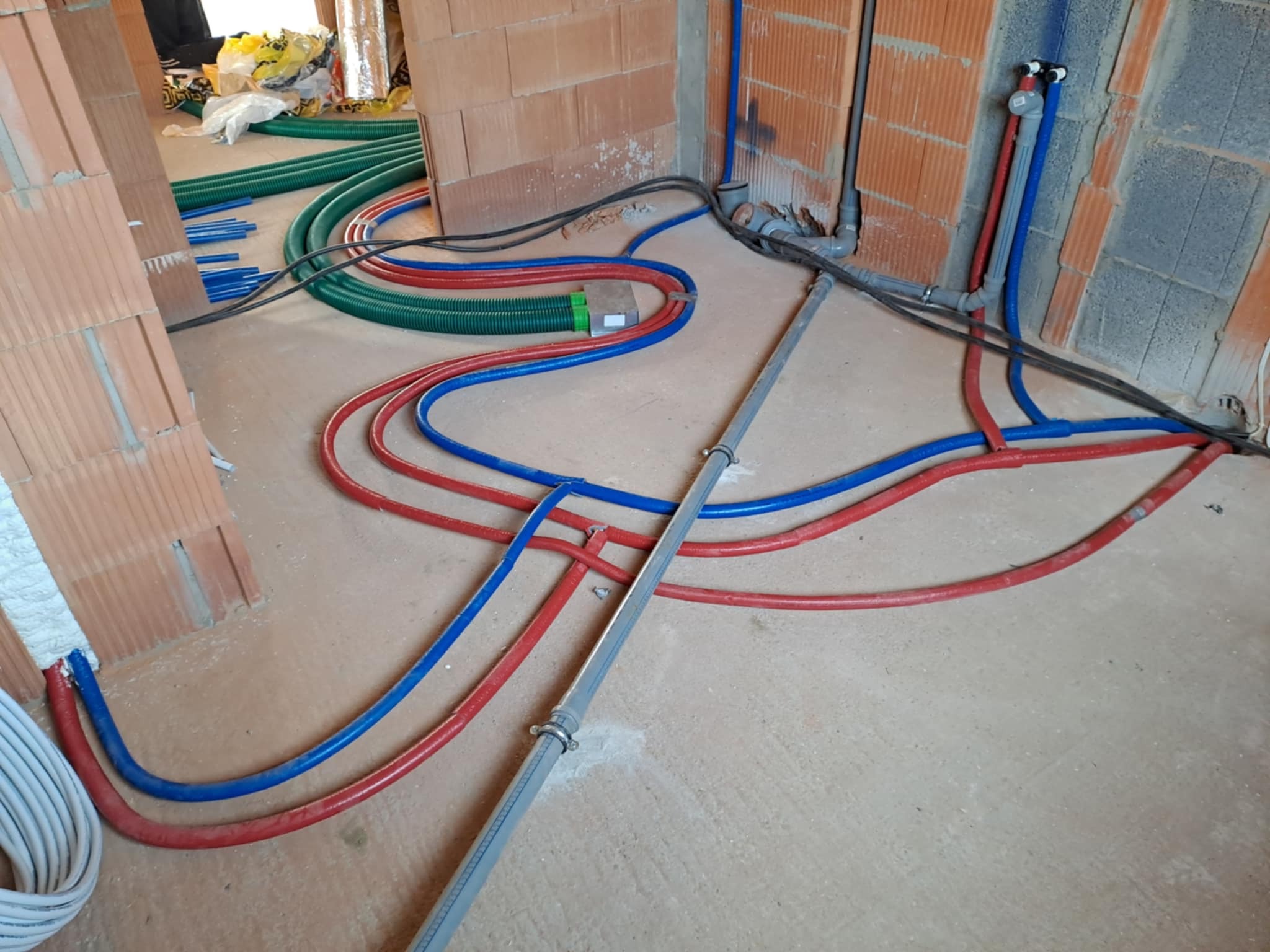 Wnętrze w trakcie prac instalacyjnych: czerwone i niebieskie rury ogrzewania podłogowego oraz zielone rury wentylacyjne ułożone na surowym podłożu, widoczne fragmenty ścian z cegły dziurawki.