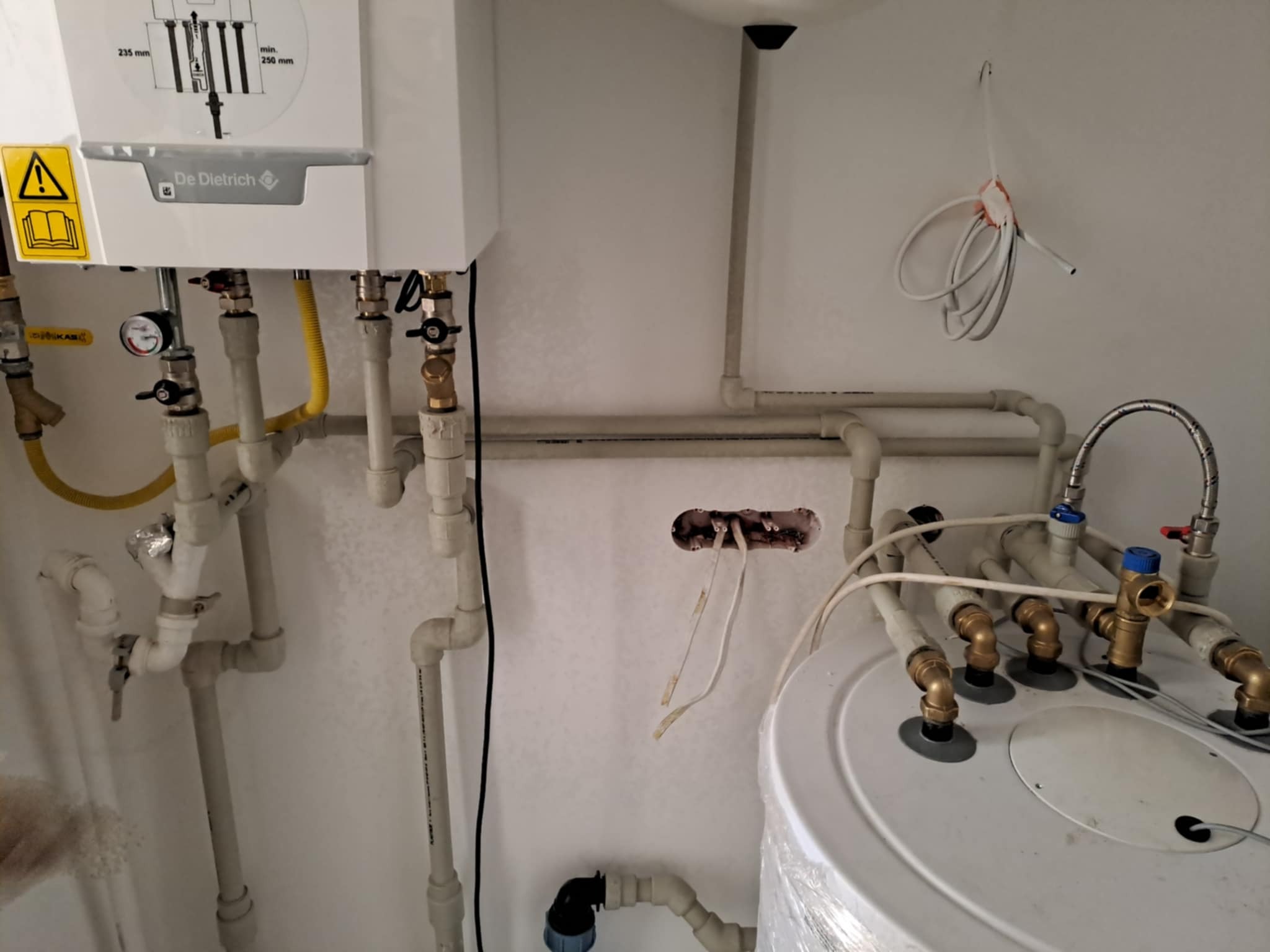 Biały piec gazowy marki De Dietrich z licznymi rurami i zaworami, widoczne elementy instalacji grzewczej i wodnej, w tle fragment ściany z wystającymi przewodami elektrycznymi.