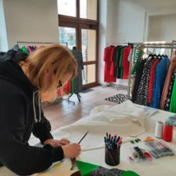 Krawcowa w czarnej bluzie z kapturem nanosi ołówkiem oznaczenia na białą tkaninę leżącą na stole, w tle wieszaki z gotowymi ubraniami.