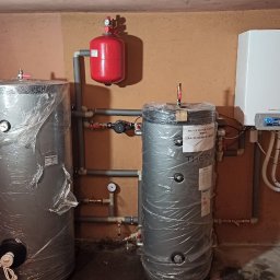 Eko Technics - Dwa srebrne zbiorniki akumulacyjne ciepła, jeden owinięty folią ochronną, połączone rurami z czerwonym zbiornikiem wyrównawczym i białym kotłem grzewczym De Dietrich na ścianie w kotłowni.