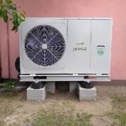 Biała jednostka zewnętrzna z wentylatorem, marki Prima, zamontowana na betonowych blokach przy ścianie budynku o różowej elewacji, widoczny fragment rynny i gałązka drzewa.