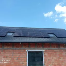 Panele fotowoltaiczne na grafitowym dachu z dwoma oknami dachowymi, zamontowane na ceglanym budynku w trakcie budowy.