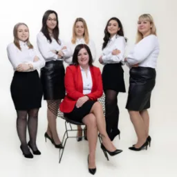 Zespół pięciu kobiet w eleganckich strojach biznesowych: czarne spódnice i białe koszule, liderka w czerwonej marynarce siedzi na krześle, z założonymi nogami, na białym tle.