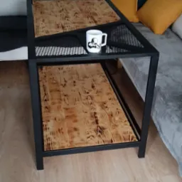 Stolik kawowy w stylu industrialnym z czarną metalową ramą i blatem z jasnego drewna o wyraźnym usłojeniu. Na g&oacute;rnej p&oacute;łce stoi biały kubek z czarnym nadrukiem. W tle widoczna sofa z poduszkami.