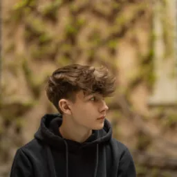 Portret młodego mężczyzny w czarnej bluzie z kapturem, profil, z kręconymi włosami na tle ściany budynku porośniętej bluszczem i okna z kratami.