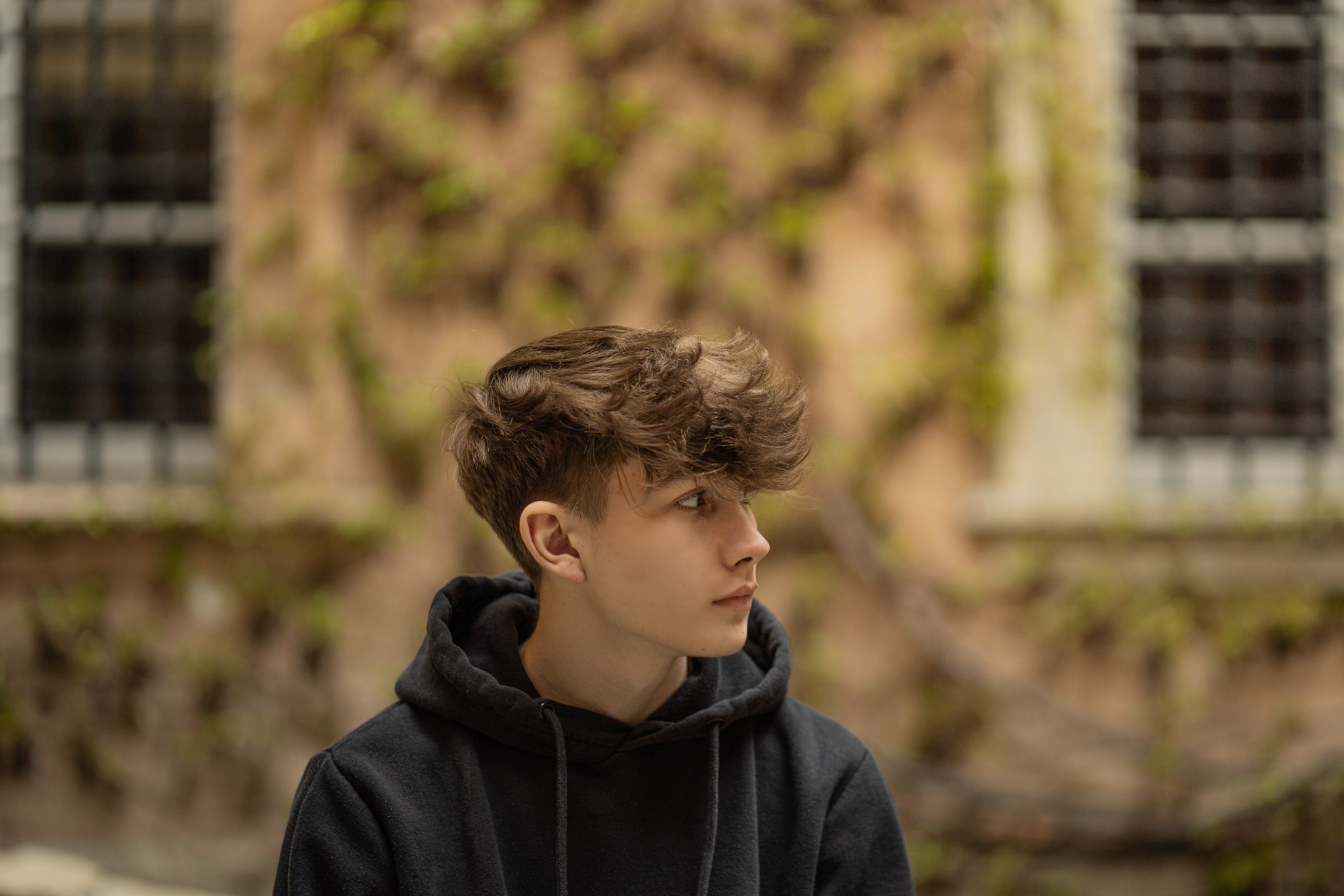 Portret młodego mężczyzny w czarnej bluzie z kapturem, profil, z kręconymi włosami na tle ściany budynku porośniętej bluszczem i okna z kratami.
