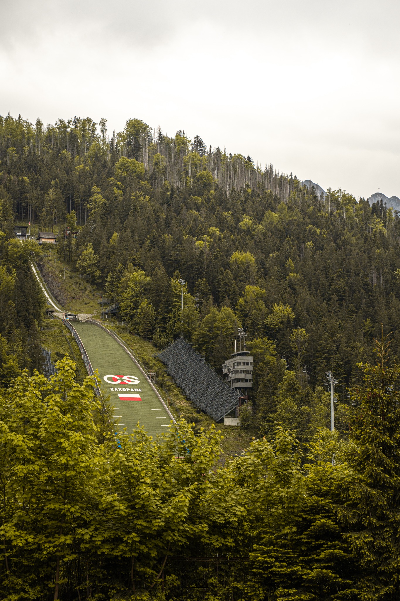 Wysoka skocznia narciarska w Zakopanem, otoczona lasem, z widoczną wieżą sędziowską i trybunami dla widzów, sfotografowana z dołu, częściowo zasłonięta przez drzewa.