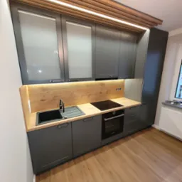 Nowoczesna kuchnia w odcieniach szarości z drewnianym blatem i oświetleniem LED pod szafkami wiszącymi, widoczna płyta indukcyjna i czarny zlew, drewniana listwa dekoracyjna na suficie.